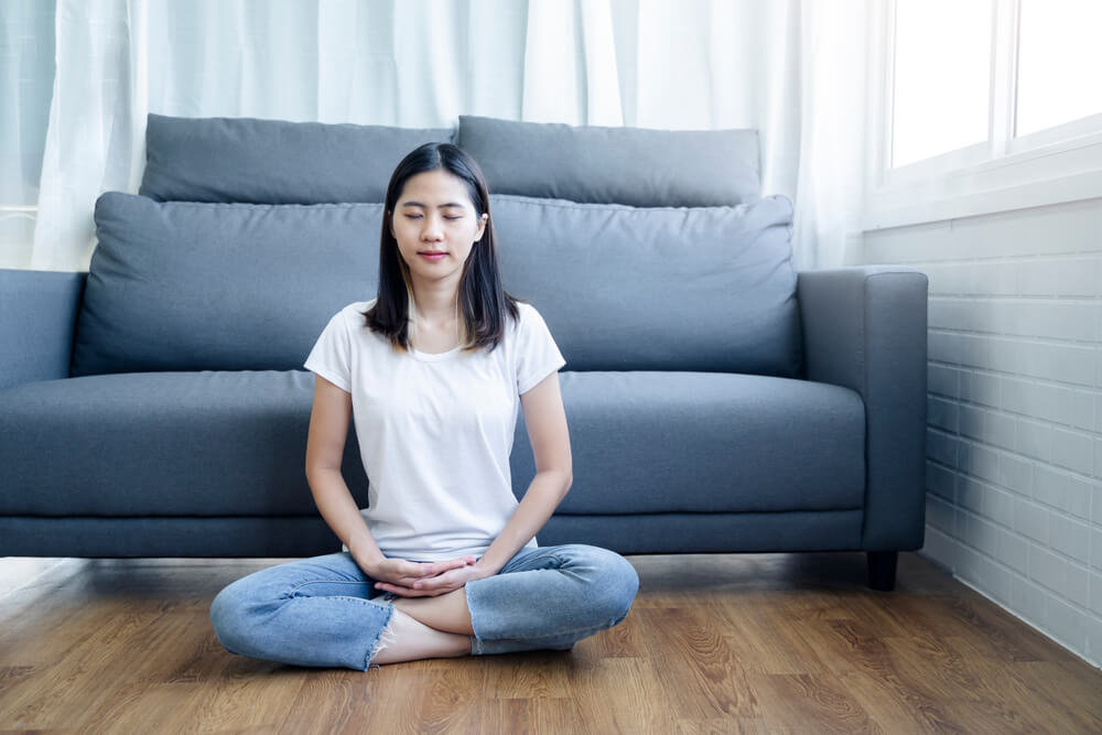 A woman practicing meditation for better sleep by a sofa.
