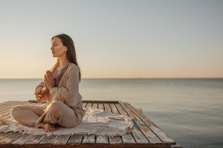 Calm young caucasian woman is meditating while sitting on wooden pier by sea.